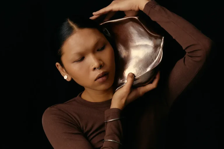 Chica que trata de escuchar hablar a una caracola que mantiene con sus manos y brazos de forma artística. Como símil de oir a la propia marca de uno.